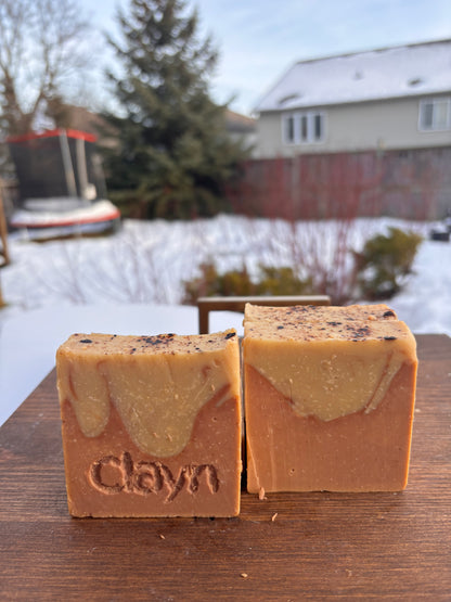 Two bars of soap with 'Claym' branding on a wooden surface outdoors with snow and trees in the background.