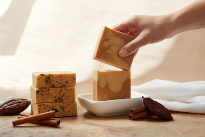 Hand holding a bar of soap with other bars stacked on a surface, surrounded by cinnamon sticks and a cloth.
